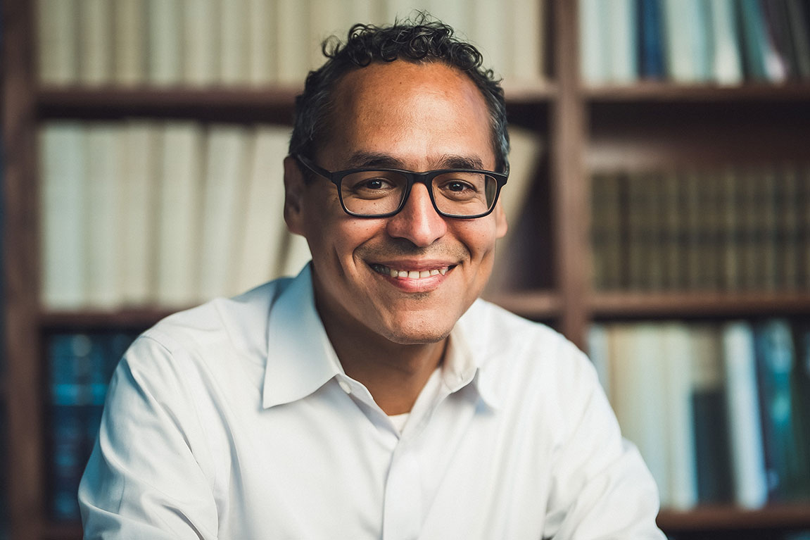 Roosevelt Montás sitting in front of library shelves of books.