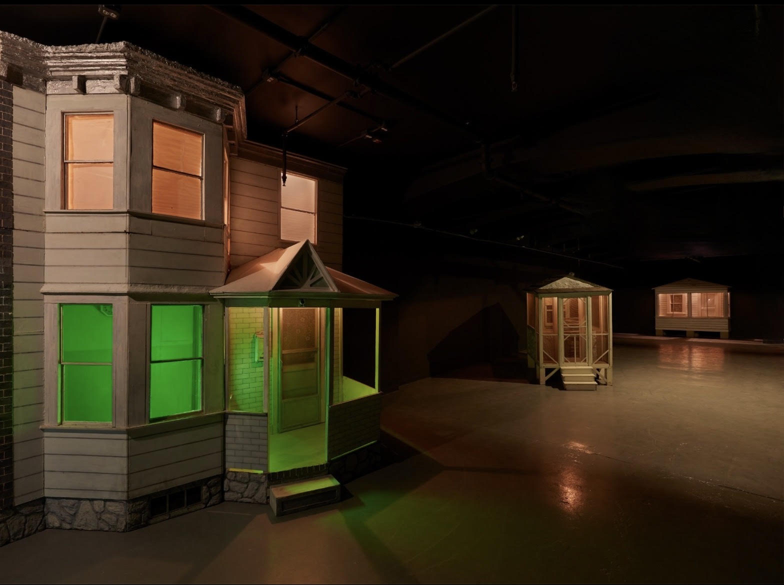 A dark room with sculptures of a facade of building in foreground with small porch and windows to the left, green light emanating from the first floor, a pinkish-orange light emanating from the top floor. The space is dark, so the light shines on the floor. In the distance is a smaller structure with the same pinkish orange light emanating from the windows.; Visit https://studioartvisitingartistdonnadennis