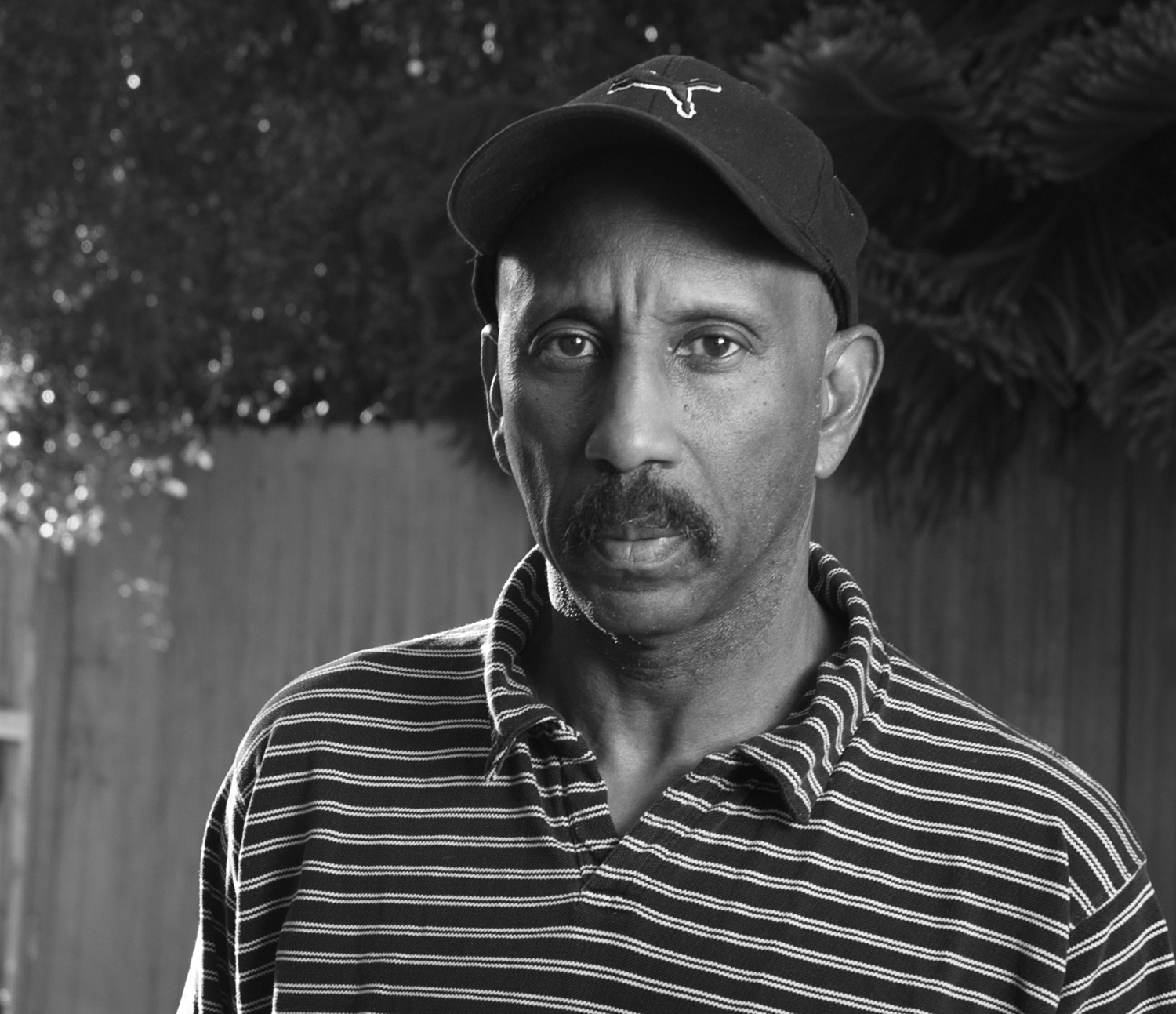 A black and white image of a man in a baseball cap looking at the camera.; A Reading with Will Alexander