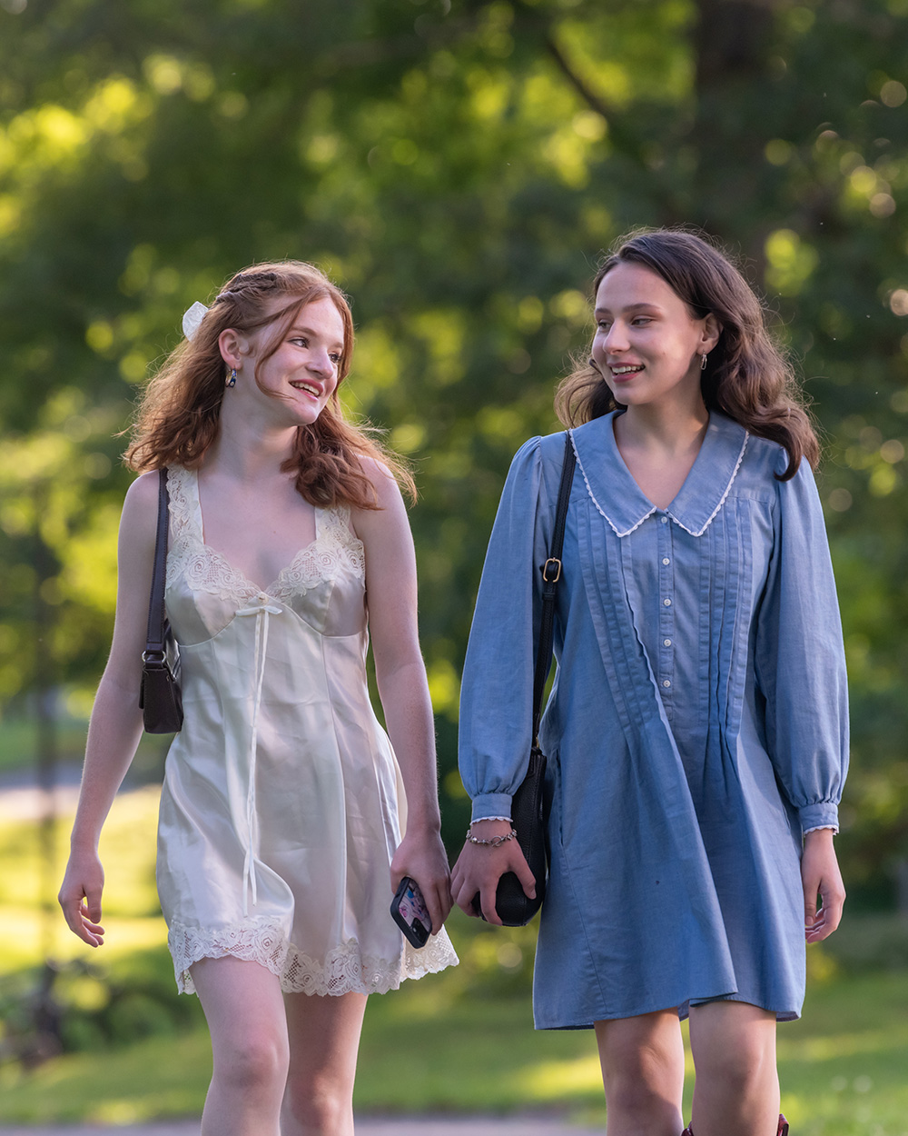 Two women walk toward the camera, smiling, with greenery behind them.