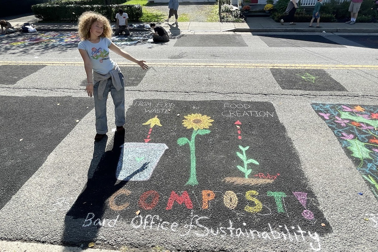 A woman poses with chalk art that reads "Compost! Bard Office of Sustainability"