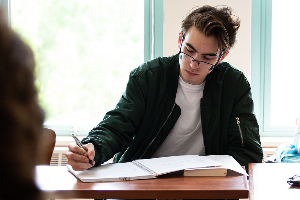 A student wearing a dark green jacket writes in a notebook.